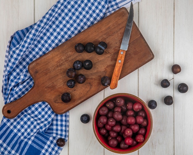 Vue de dessus des petites prunelles bleu-noir aigre sur une planche de cuisine en bois avec un couteau avec des cerises rouges sur un bol rouge sur un fond en bois gris