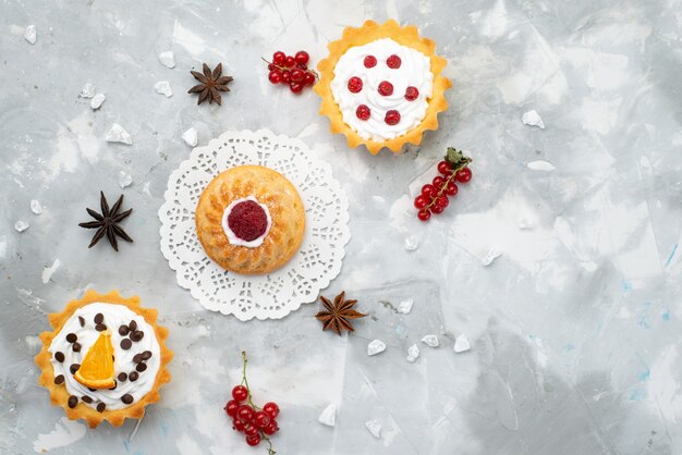 Vue de dessus petits gâteaux délicieux avec de la crème et des fruits rouges sur le gâteau de bureau gris crème au sucre biscuit sucré