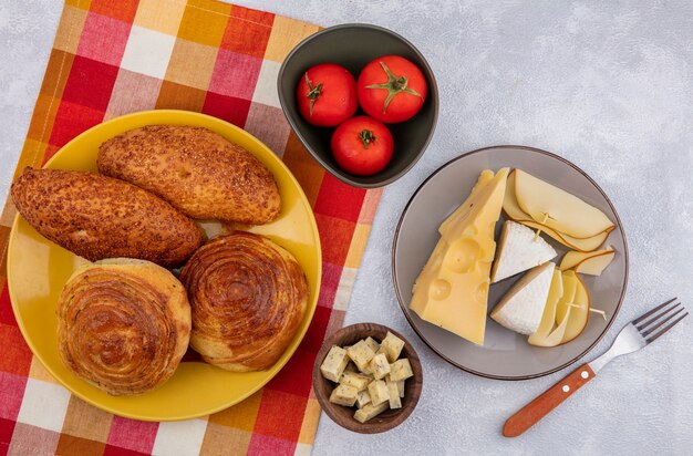 Vue de dessus de petits pains frais sur une plaque jaune sur un chiffon vérifié avec différents types de fromage sur une plaque grise avec des tomates sur un bol sur un fond blanc