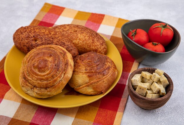 Vue de dessus des petits pains sur une plaque jaune sur un chiffon vérifié avec du fromage sur un bol en bois avec des tomates sur un bol sur un fond blanc