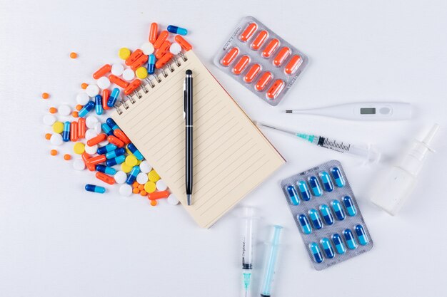Vue de dessus des pilules colorées avec bloc-notes, stylo, thermomètre, vaporisateur nasal et aiguille