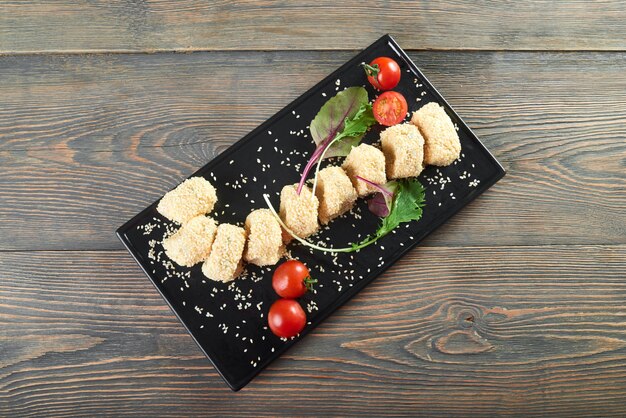 Vue de dessus plan horizontal d'une palette noire avec de délicieux rouleaux de fromage avec du sezam servi avec des tomates cerises et quelques feuilles de table en bois verts