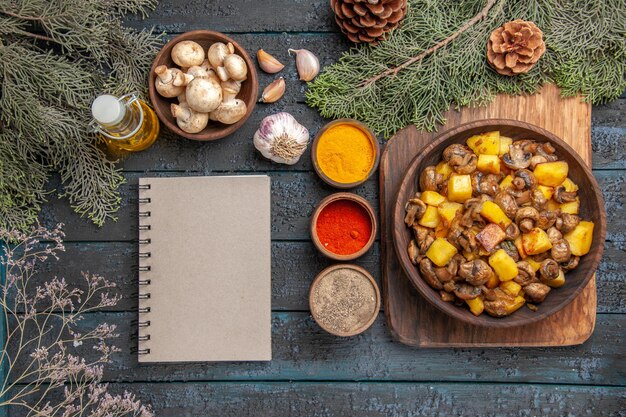 Vue de dessus plat et assiette de champignons et de pommes de terre sur une planche à découper à côté d'huile de cahier d'épices colorées en bouteille bol d'ail de champignons et de branches avec des cônes