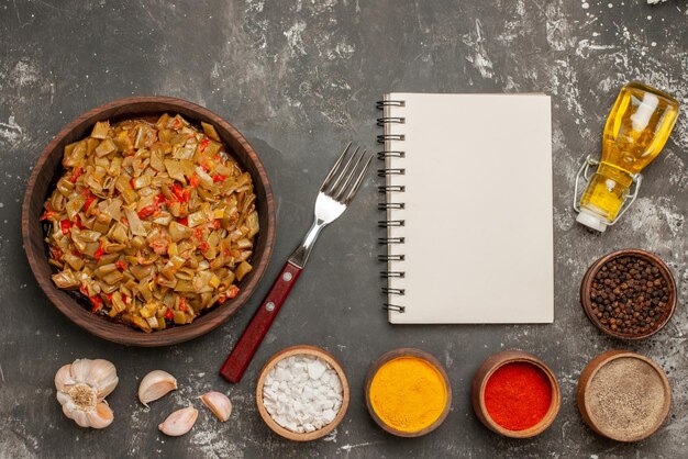 Vue de dessus plat savoureux les haricots verts appétissants avec des tomates à côté des bols de cahier blanc de cinq sortes d'épices fourchette à l'ail bouteille d'huile sur la table sombre