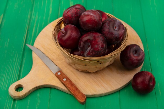 Photo gratuite vue de dessus des pluots frais sur un seau sur une planche de cuisine en bois avec un couteau sur un mur en bois vert