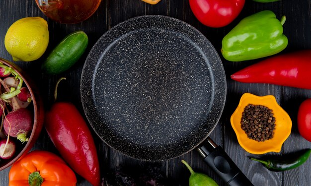 Vue de dessus de la poêle et des légumes frais poivrons colorés tomates radis concombres et grains de poivre noir disposés autour sur une table en bois foncé