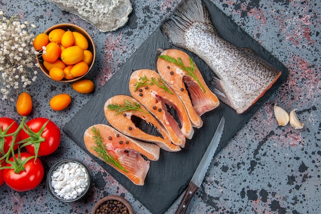 Vue de dessus des poissons crus frais verts sur plateau de couleur sombre épices tomates avec tiges ail fleurs couteau kumquats sur table de couleurs de mélange noir bleu