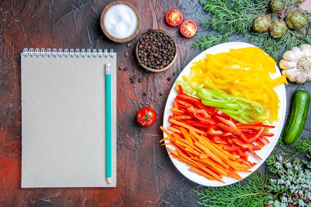 Vue de dessus poivrons coupés colorés sur plaque sel et poivre noir tomates ail concombre crayon sur bloc-notes sur table rouge foncé