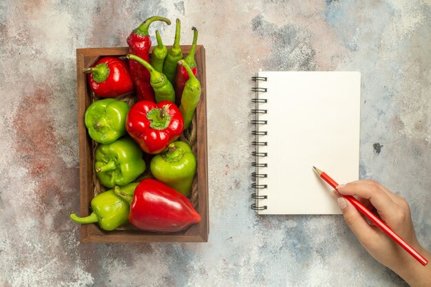 Vue de dessus poivrons rouges et verts piments dans une boîte en bois un crayon de cahier en main de femme sur une surface nue