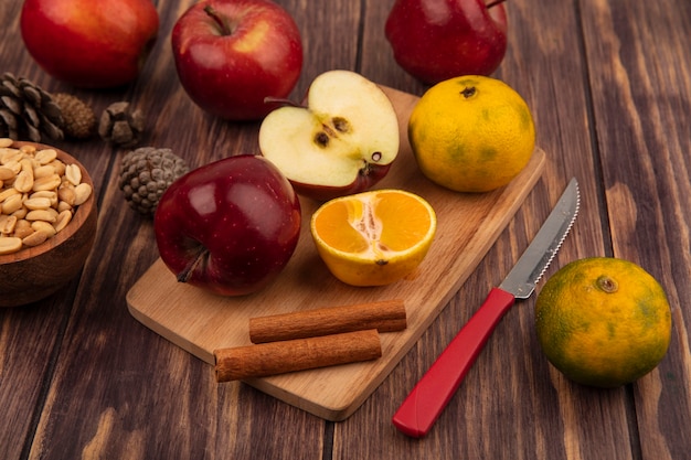 Vue de dessus des pommes biologiques sur une planche de cuisine en bois avec la moitié des mandarines et des bâtons de cannelle avec un couteau avec des arachides sur un bol en bois avec des pommes isolé sur un fond en bois
