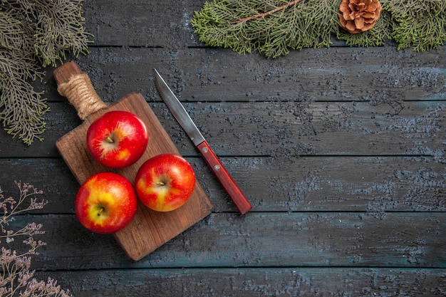 Vue de dessus des pommes à bord de trois pommes jaune-rougeâtre sur la planche à découper brune sous les arbres avec des cônes sur le côté gauche de la table