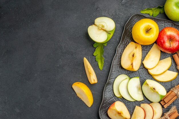 Vue de dessus des pommes fraîches tranchées et entières vertes, jaunes et rouges sur un plateau noir et des citrons verts à la cannelle sur le côté gauche sur une table sombre