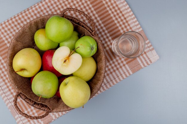 Vue de dessus des pommes rouges jaunes vertes dans le panier et verre d'eau sur tissu à carreaux et fond gris