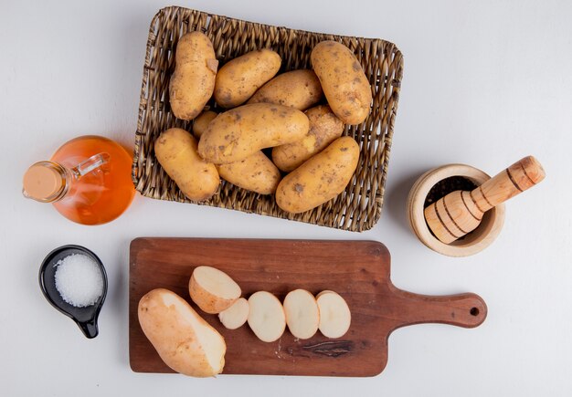 Vue de dessus des pommes de terre coupées et tranchées sur une planche à découper avec d'autres dans l'assiette du panier avec du sel poivre noir beurre fondu sur la surface blanche