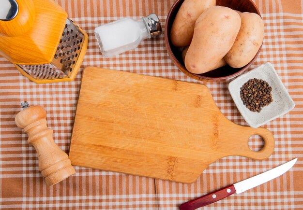 Vue de dessus des pommes de terre dans un bol avec un couteau à râper le sel au poivre noir autour d'une planche à découper sur fond de tissu à carreaux