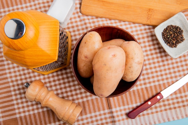 Vue de dessus des pommes de terre dans un bol avec un couteau à râper le sel au poivre noir et une planche à découper sur fond de tissu à carreaux