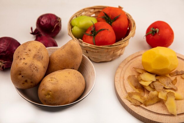 Vue de dessus de pommes de terre pelées bio sur une planche de cuisine en bois avec des pommes de terre sur un bol avec des tomates et du poivre sur un seau avec des oignons rouges isolé sur une surface blanche