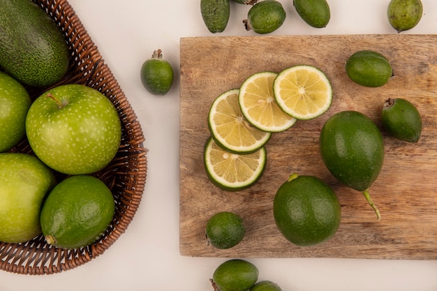 Vue de dessus des pommes vertes avec des avocats sur un seau avec des limes et des feijoas sur une planche de cuisine en bois sur un mur blanc