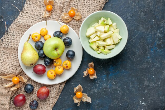 Vue de dessus des pommes vertes moelleuses et juteuses avec des tranches de pomme à l'intérieur de la plaque avec d'autres fruits sur un bureau bleu foncé, fruits frais santé vitamine