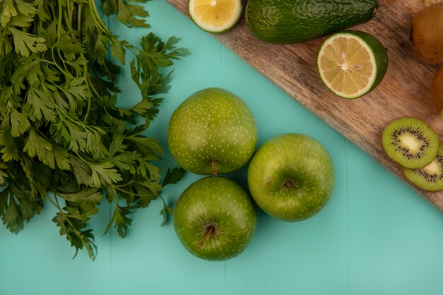 Vue de dessus des pommes vertes saines avec des avocats limes et des kiwis sur une planche de cuisine en bois sur un mur bleu
