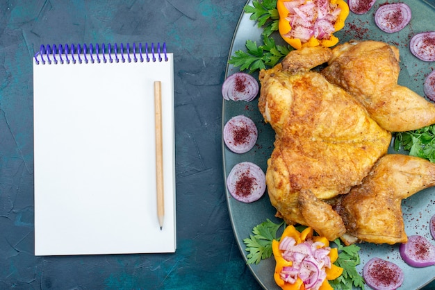 Vue de dessus poulet cuit avec des oignons et des légumes verts à l'intérieur de la plaque avec bloc-notes sur le bureau bleu foncé viande de poulet cuire le dîner au four