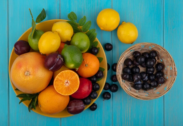 Vue de dessus prune cerise dans un panier avec des oranges prunes citrons au citron vert sur une plaque jaune sur fond turquoise