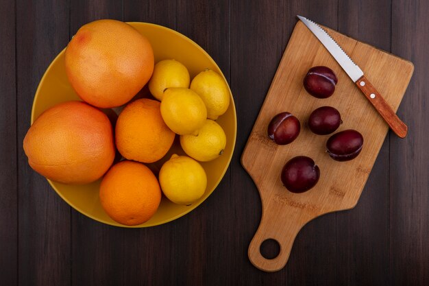 Vue de dessus prune sur une planche à découper avec un couteau avec des citrons, des oranges et des pamplemousses dans un bol sur un fond en bois