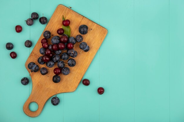 Vue de dessus des prunelles violet foncé avec des cerises sur une planche de cuisine en bois sur fond bleu avec espace copie