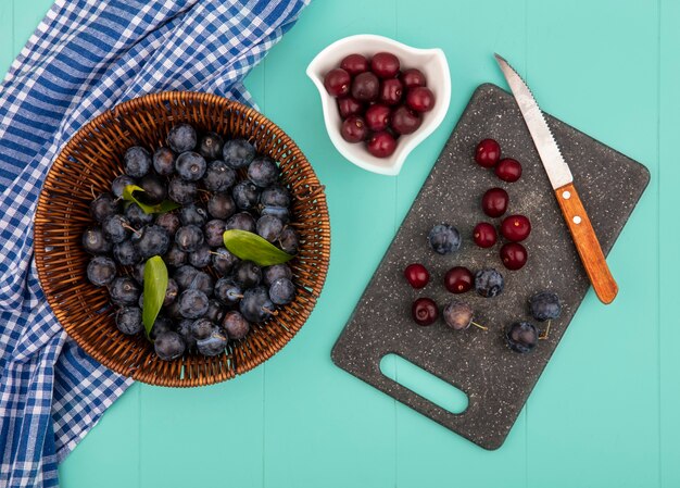 Vue de dessus des prunelles violet foncé sur un seau avec de délicieuses cerises rouges sur un bol blanc avec des prunelles et des cerises sur une planche à découper de cuisine avec un couteau sur un fond bleu