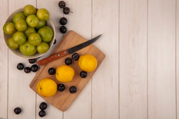 Vue de dessus des prunes cerises vertes sur un bol avec des pêches jaunes sur une planche de cuisine en bois avec un couteau avec des prunes sur un fond en bois gris avec espace copie