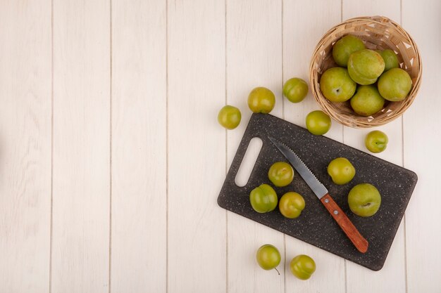 Vue de dessus des prunes cerises vertes fraîches sur une planche à découper de cuisine avec un couteau avec des prunes cerises sur un seau sur un fond en bois blanc avec espace de copie