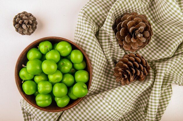 Photo gratuite vue de dessus des prunes vertes aigres dans un bol en bois et des cônes sur tissu écossais
