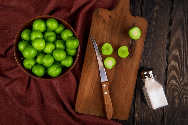 Photo gratuite vue de dessus des prunes vertes aigres dans un bol et des prunes en tranches sur une planche à découper en bois avec un couteau et une salière sur une table rustique sombre