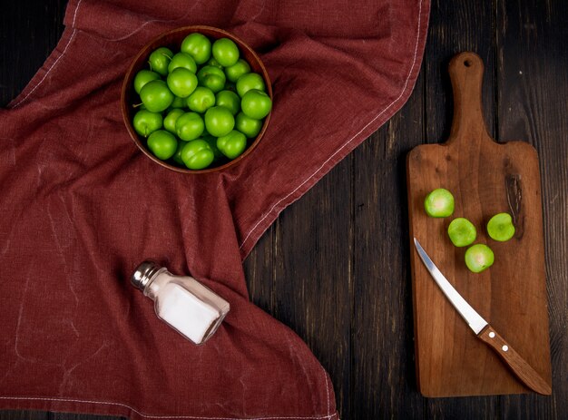 Vue de dessus des prunes vertes aigres dans un bol et des prunes en tranches sur une planche à découper en bois avec un couteau et une salière sur une table rustique sombre