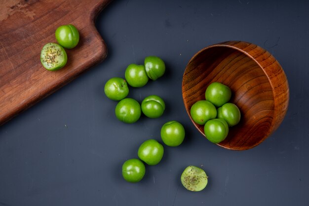 Vue de dessus des prunes vertes aigres éparpillées dans un bol en bois et une planche à découper en bois avec des prunes en tranches sur un tableau noir