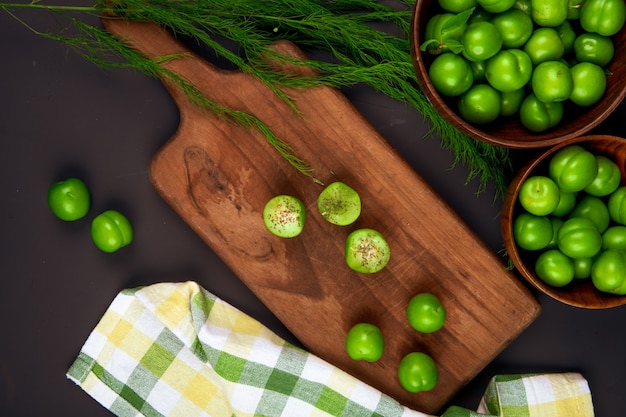Vue de dessus de prunes vertes tranchées saupoudrées de menthe poivrée séchée sur une planche à découper en bois et des bols en bois remplis de prunes vertes sur tableau noir