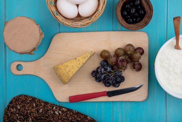 Vue de dessus des raisins avec du fromage sur un stand avec des olives oeufs de poule yaourt et pain noir sur fond turquoise