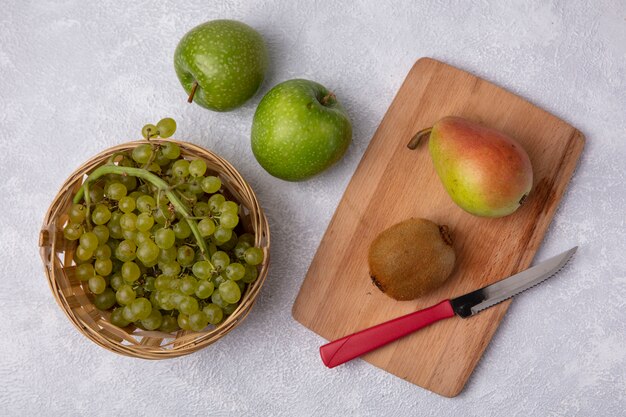 Vue de dessus des raisins verts dans un panier avec kiwi poire et couteau sur une planche à découper avec des pommes vertes sur fond blanc