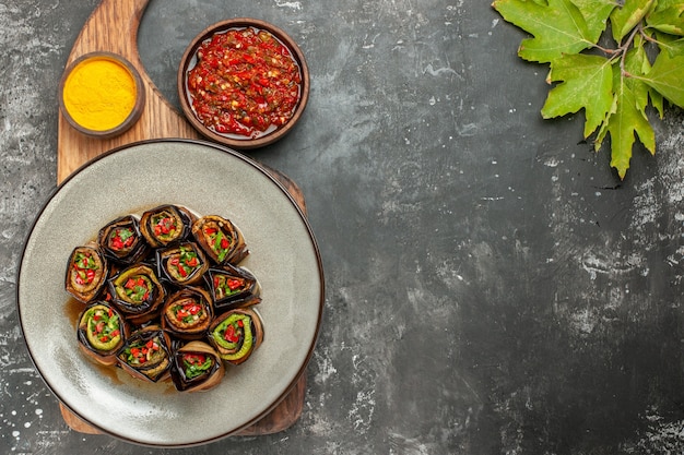 Vue de dessus des rouleaux d'aubergine farcis dans une assiette ovale blanche au curcuma dans un bol sur un plateau de service en bois adjika sur fond gris