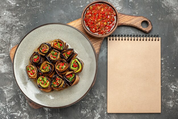 Vue de dessus des rouleaux d'aubergine farcis dans une assiette ovale blanche poivre noir dans un bol sur une planche de service en bois avec poignée adjika un cahier sur une surface grise