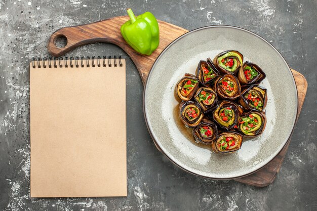 Vue de dessus des rouleaux d'aubergine farcis dans une assiette ovale un poivron vert sur une planche de service en bois avec poignée différentes épices dans de petits braillements un cahier sur une surface grise