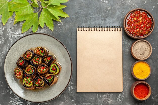 Vue de dessus des rouleaux d'aubergines farcies dans une assiette blanche différentes épices adjika dans de petits bols un cahier sur une surface grise