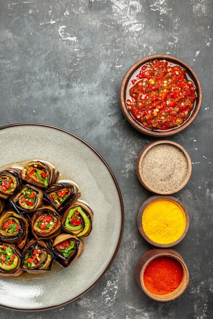 Vue de dessus des rouleaux d'aubergines farcies dans une assiette blanche différentes épices adjika dans de petits bols sur une surface grise