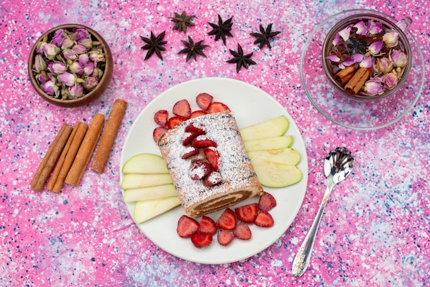 Vue de dessus rouler le gâteau à l'intérieur de la plaque avec des pommes et des fraises avec de la cannelle et du thé sur le gâteau de fond coloré cuire des fruits sucrés