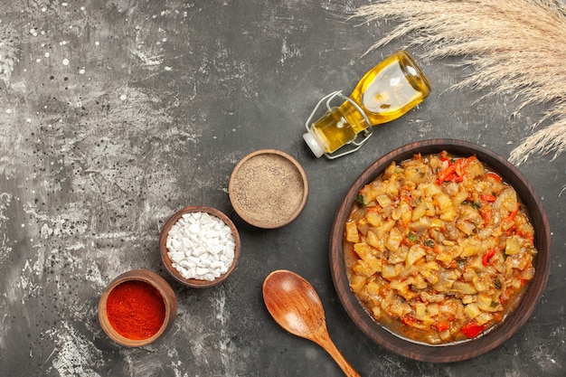 Vue de dessus de la salade d'aubergines rôties dans un bol, une bouteille d'huile, une cuillère en bois et différentes épices dans des bols sur une surface sombre