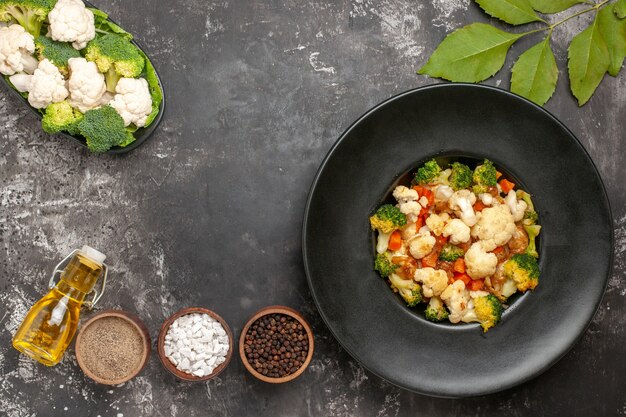 Vue de dessus salade de brocoli et de chou-fleur dans un bol noir différentes épices dans des bols de légumes crus d'huile sur une plaque sur une surface sombre