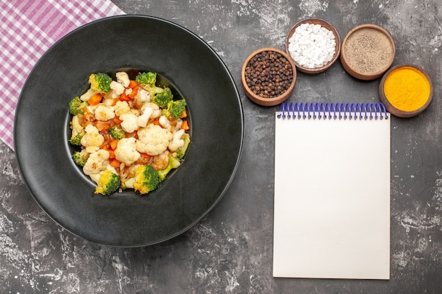 Vue de dessus salade de brocoli et de chou-fleur dans un bol noir serviette à carreaux rose et blanc différentes épices un cahier sur une surface sombre