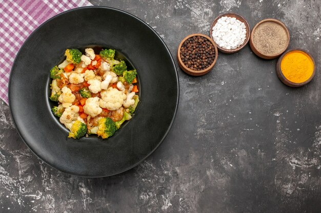 Vue de dessus salade de brocoli et de chou-fleur dans un bol noir serviette à carreaux rose et blanc différentes épices sur espace libre surface sombre