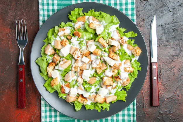 Vue de dessus salade césar sur assiette ovale nappe à carreaux blanc vert fourchette et couteau sur fond rouge foncé