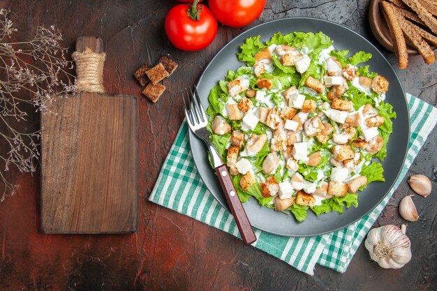 Vue de dessus salade césar sur assiette ovale serviette à carreaux blanc vert planche à découper ail sur fond rouge foncé